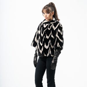 Puede incluir: Chaqueta de piel sintética en blanco y negro con estampado de chevrón. La chaqueta tiene un cuello ancho y mangas cortas. La modelo lleva guantes negros y pantalones negros. La chaqueta es una prenda de abrigo elegante.
