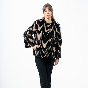 May include: A black and tan faux fur jacket with a chevron pattern. The jacket has long sleeves and is paired with black pants and black heels. The model is standing in front of a white background.