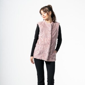 Könnte beinhalten: Eine hellrosa Kunstfellweste mit strukturiertem Muster. Die Weste wird über einem schwarzen Langarmshirt getragen und mit einer schwarzen Hose kombiniert. Das Model steht vor einem weißen Hintergrund.