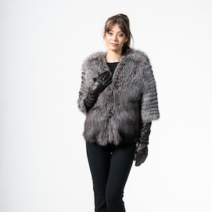 Könnte beinhalten: Silberfuchs-Pelzjacke mit schwarzem Futter und schwarzen Lederhandschuhen. Die Jacke hat ein kurzes Design mit Dreiviertelärmeln. Die Person trägt eine schwarze Hose und schwarze Absätze. Der Hintergrund ist weiß.
