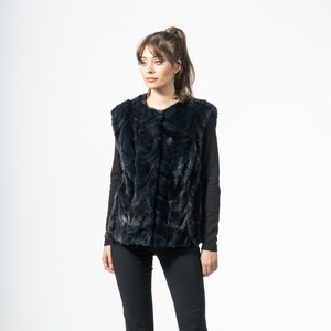 May include: A dark blue faux fur vest with a textured pattern. The vest is worn over a black long-sleeve shirt and paired with black pants. The outfit is completed with black pointed-toe heels.