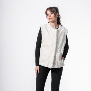 Puede incluir: Un chaleco de piel sintética color crema con un patrón texturizado. El chaleco se lleva sobre una camiseta negra de manga larga y se combina con pantalones negros. El atuendo se completa con tacones negros. El estilo general es elegante y chic.