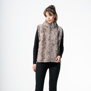 May include: A woman wearing a taupe faux fur vest with a high collar and a full-length zipper. She is also wearing a black long-sleeve shirt and black pants. The vest has a textured pattern.