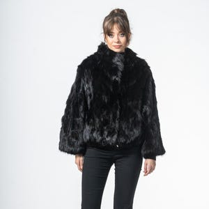 Puede incluir: Chaqueta de piel negra con cuello alto y mangas acampanadas. La chaqueta se combina con pantalones negros y zapatos de tacón negros. La mujer está de pie frente a un fondo blanco.