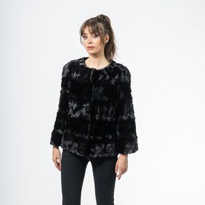 May include: A black faux fur jacket with a round collar and long sleeves. The jacket is paired with black pants and black heels. The woman is standing in front of a white background.
