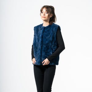 Puede incluir: Una mujer con un chaleco de piel sintética azul oscuro sobre una camisa negra de manga larga y pantalones negros. El chaleco tiene una apariencia texturizada. La mujer está de pie frente a un fondo blanco.