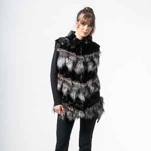 May include: A black and silver faux fur vest with a layered design. The vest is worn over a black long-sleeved top, paired with black trousers and heels. The vest's fur has a mix of black and silver tones.