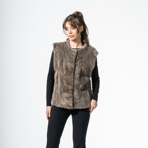 May include: A brown fur vest with a textured appearance. The vest is worn over a black long-sleeve top and paired with black pants and black heels. The outfit is set against a white background.