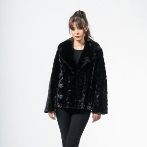 May include: A black faux fur jacket with a wide collar and a textured appearance. The jacket is worn over a dark top and paired with black pants and heels. The overall look is elegant and stylish.