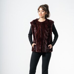 May include: A burgundy fur vest with a round neckline. The vest is worn over a black long-sleeve top and paired with black pants. The model is wearing black high heels. The background is white.
