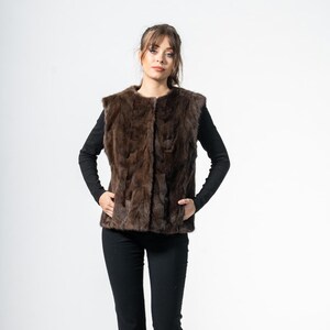 May include: A brown fur vest with a textured appearance. The vest is worn over a black long-sleeve shirt and paired with black pants. The model is standing in front of a white background.