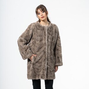 May include: A long, light brown fur coat with a textured pattern. The coat has long sleeves and a round neckline. The person in the image is wearing black pants and black shoes. The background is white.