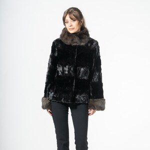 Puede incluir: Una mujer con una chaqueta de piel negra con cuello y puños de piel marrón, combinada con pantalones negros y tacones negros. La chaqueta tiene un aspecto texturizado. El fondo es blanco liso.