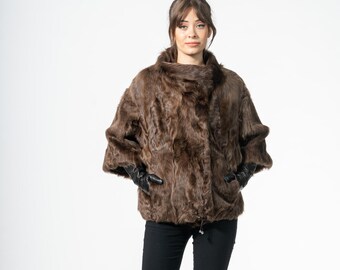 May include: A brown fur coat with a high collar and short sleeves. The coat is paired with black pants and black gloves. The model is standing in front of a white background. The coat appears to be made of a soft, luxurious material.