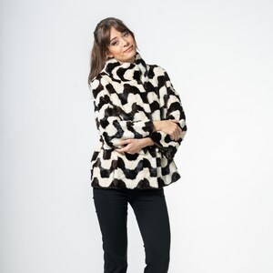Puede incluir: Chaqueta de piel sintética con estampado de zigzag en blanco y negro y cuello alto. La persona lleva pantalones negros y tacones negros. El fondo es blanco. Prenda de abrigo elegante.