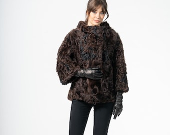 May include: A brown fur jacket with a textured appearance. The jacket has a rounded collar and three-quarter sleeves. The person is wearing black gloves and black pants. The pants have a slight flare at the bottom.