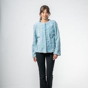 Puede incluir: Chaqueta de piel sintética azul claro con cuello redondo y mangas largas. La chaqueta se usa con pantalones negros y zapatos negros. La chaqueta tiene una apariencia texturizada y un aspecto suave y afelpado. El conjunto está sobre un fondo blanco.