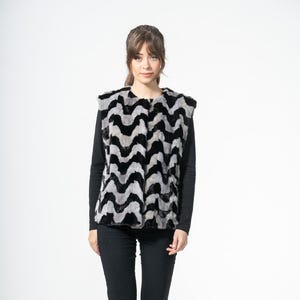 May include: A sleeveless vest with a black, gray, and white zigzag pattern. The vest is worn over a black long-sleeve shirt and paired with black pants. The model is wearing black shoes.