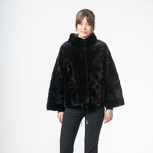 Puede incluir: Chaqueta negra de piel sintética con cuello alto y cintura con cordón. La chaqueta se combina con pantalones negros y tacones negros de punta. El conjunto se presenta sobre un fondo blanco.