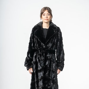 May include: A full-length black faux fur coat with a shawl collar and a matching belt. The coat has long sleeves and is worn over black pants and shoes. The coat is a luxurious outerwear garment.