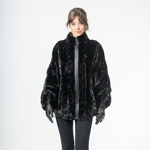 May include: A black faux fur jacket with a high collar and a zipper closure. The jacket has long sleeves and is paired with black pants and black gloves. The model is wearing black heels.