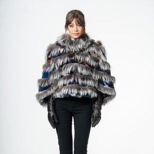 Puede incluir: Chaqueta de piel multicolor con diseño en capas. La chaqueta presenta tonos de gris, azul y rojo. La modelo lleva pantalones negros y guantes negros. El fondo es blanco.