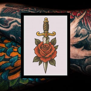 Könnte beinhalten: Gerahmter Druck eines traditionellen Tattoo-Designs. Es zeigt eine rote Rose mit grünen Blättern, durchbohrt von einem Dolch mit braunem Griff und goldenen Details. Der Hintergrund ist ein hellrosa Gitter.