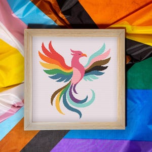 May include: A framed cross-stitch of a colorful phoenix with outstretched wings. The bird features a rainbow of colors, including red, orange, yellow, green, blue, and purple. The frame is made of light-colored wood. The background is a colorful fabric.
