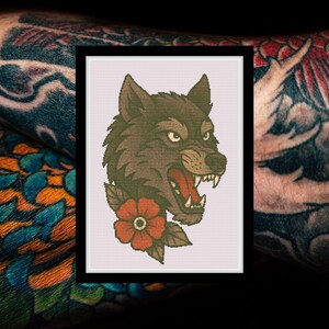 May include: A cross stitch pattern featuring a snarling grey wolf head with sharp teeth and an open mouth, accented by a single red flower with green leaves at the base. The design is presented on a white grid background within a black frame.