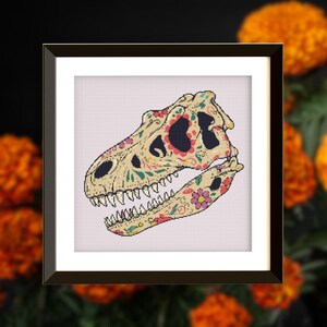 Pode incluir: Arte em ponto cruz emoldurada com um crânio de dinossauro decorado com padrões florais coloridos. O crânio é bege com detalhes em rosa, verde e amarelo. A obra de arte está em um fundo branco e emoldurada em uma moldura marrom escuro.