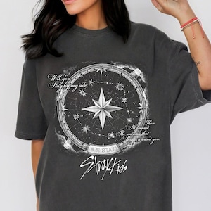 Peut inclure: T-shirt gris fonc&eacute; avec un motif de boussole, une &eacute;toile centrale et le texte "Will you stay by my side, then the universe that wraps around you." Le t-shirt porte &eacute;galement l'inscription "Stray Kids".