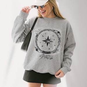 Peut inclure: Un sweat-shirt gris avec un motif de boussole, une &eacute;toile centrale et le texte "Stray Kids" en noir et blanc. Le sweat-shirt a des manches longues et un col rond. Le design est circulaire.