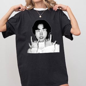 以下が含まれることがあります： 「ROCKSTAR」の文字が額に書かれた人物の白黒グラフィックが特徴の黒いTシャツ。その人物はハンドジェスチャーをしています。このシャツはクルーネックで半袖です。