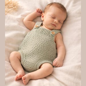 Może przedstawiać: Śpiące dziecko w jasnozielonym dzianinowym śpioszku z drewnianymi guzikami. Dziecko leży na miękkim, kremowym kocu. Śpioch to jednoczęściowy element garderoby o teksturowanym wzorze.