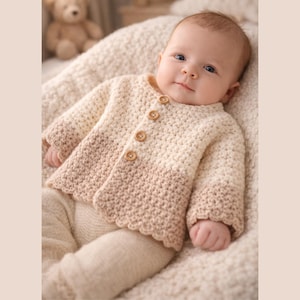 May include: A baby wearing a cream-coloured, crocheted cardigan with three wooden buttons. The cardigan has a scalloped edge and a gradient colour from cream to light brown. The baby is also wearing cream-coloured trousers.