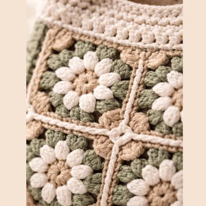 Puede incluir: Primer plano de un bolso de ganchillo con un estampado floral. El bolso presenta un dise&ntilde;o de cuadrados de la abuela en tonos crema, verde y marr&oacute;n. Las puntadas de ganchillo son visibles, creando una superficie texturizada. El borde superior del bolso es de color crema liso.