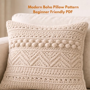 May include: A beige, crocheted pillow with a modern boho design. The pillow features various textured patterns, including bobbles and chevron shapes. Text on the image reads "Modern Boho Pillow Pattern Beginner Friendly PDF". The pillow is placed on a beige sofa.