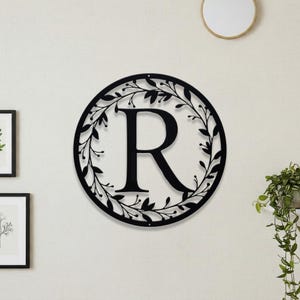 May include: A black metal wall decoration with the letter "R" at its centre, encircled by a wreath of leaves. The circular design is mounted on a white wall, with other framed artwork and a mirror visible in the background.