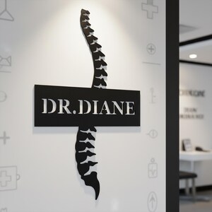 May include: A black and white sign for a medical office. The sign features the text "DR. DIANE" in white, with a stylized spine design above and below the text. The sign is mounted on a white wall.