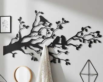 Puede incluir: Arte mural de metal negro que representa una rama de árbol con hojas y dos pájaros posados. La rama tiene ganchos para colgar objetos. Una manta blanca cuelga de uno de los ganchos. Hay objetos decorativos en la pared.