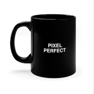 Może przedstawiać: Błyszczący czarny kubek ceramiczny z zakrzywionym uchwytem i napisem PIXEL PERFECT wydrukowanym pogrubionymi białymi wielkimi literami z przodu. Kubek ma gładkie, jednolite wykończenie oraz standardowy cylindryczny kształt z szeroką krawędzią i podstawą.