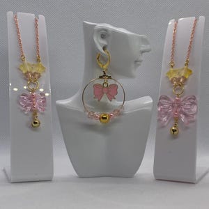 Pode incluir: Um conjunto de joias rosa e douradas, incluindo dois colares e um par de brincos de argola. Os colares apresentam um pingente de laço rosa e um berloque de sino dourado. Os brincos têm um laço rosa e contas rosas.
