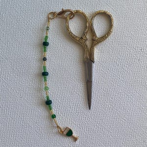 Può includere: Forbici da ricamo dorate con un ciondolo di perline. Il ciondolo presenta perline verdi e dorate e un piccolo calice dorato con smalto verde. Le forbici hanno manici ornati e lame argentate.