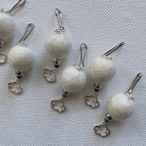 Może przedstawiać: Kilka białych filcowych zawieszek w kształcie kuli ze srebrnymi zapięciami i zawieszkami w kształcie chmur. Każda zawieszka ma małą niebieską koralik i srebrną zawieszkę w kształcie chmury.