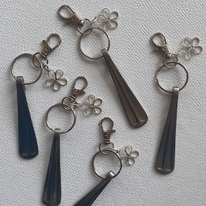 May include: A collection of keychains with a metal spoon design. Each keychain has a silver-colored clasp, a large ring, and a decorative flower charm. The spoons are dark gray and have a sleek, elongated shape.