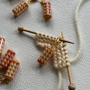 Op de afbeelding: Close-up van breinaalden met een klein gebreid lapje en verschillende oranje en witte geometrische steekmarkeerders. De steekmarkeerders zijn cilindrisch met goudkleurige sluitingen. Crèmekleurig garen is ook zichtbaar.