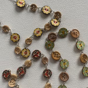 May include: A handcrafted necklace composed of wooden buttons in various sizes and designs. Some buttons display floral patterns in yellow, red, and green. The necklace is strung on a silver chain, offering a distinctive, artistic accessory.