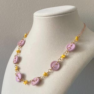 May include: A necklace featuring lavender-colored button-shaped beads and yellow star-shaped charms. The beads and charms are connected by a delicate gold-colored chain. The necklace is displayed on a white mannequin bust.