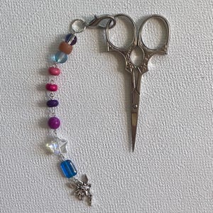 Può includere: Forbici da ricamo argentate con un ciondolo di perline. Il ciondolo presenta perline rosa, viola, blu e marroni, un ciondolo a forma di fata argentata e una perlina a forma di stella. Per cucito.
