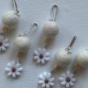 May include: Several handmade daisy earrings with a white felt ball, wooden bead, and silver-tone clasp. The daisy flowers have white petals and a brown center. The earrings are displayed on a textured white surface.